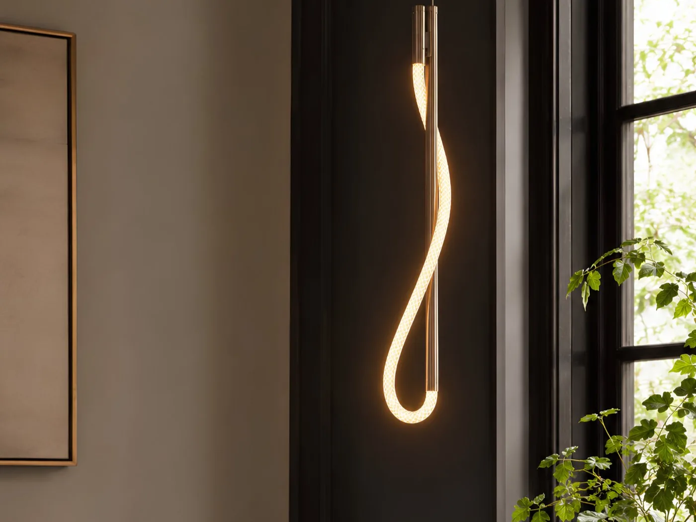 Eden Pendant in polished nickel — a single illuminated cord looped around a slim vertical stem, against dark paneling beside a window