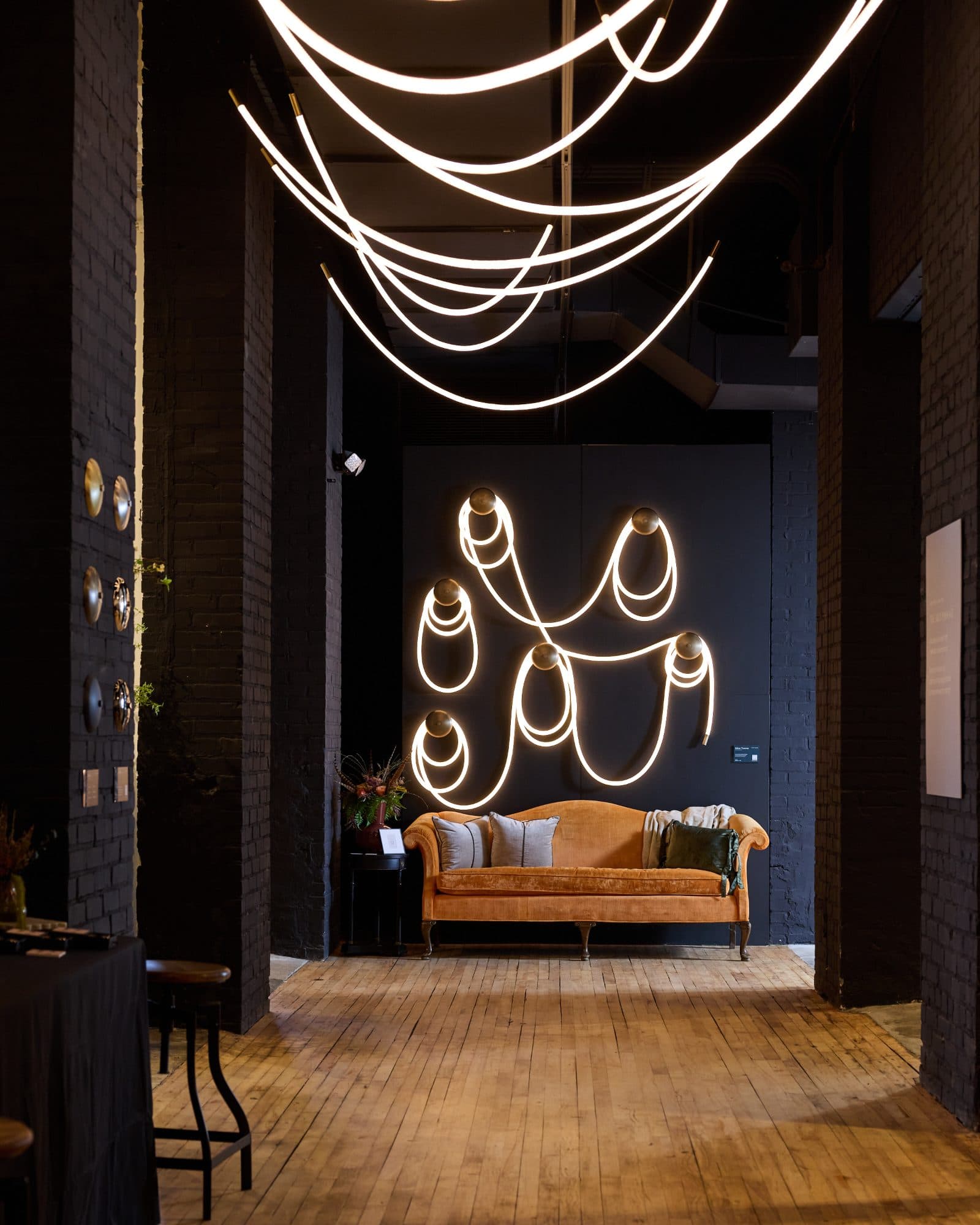 Selene Traverse on a dark wall at High Point Market, Fall 2026: illuminated braided cord looping between six wall canopies above a velvet sofa, with illuminated cord pendants overhead.